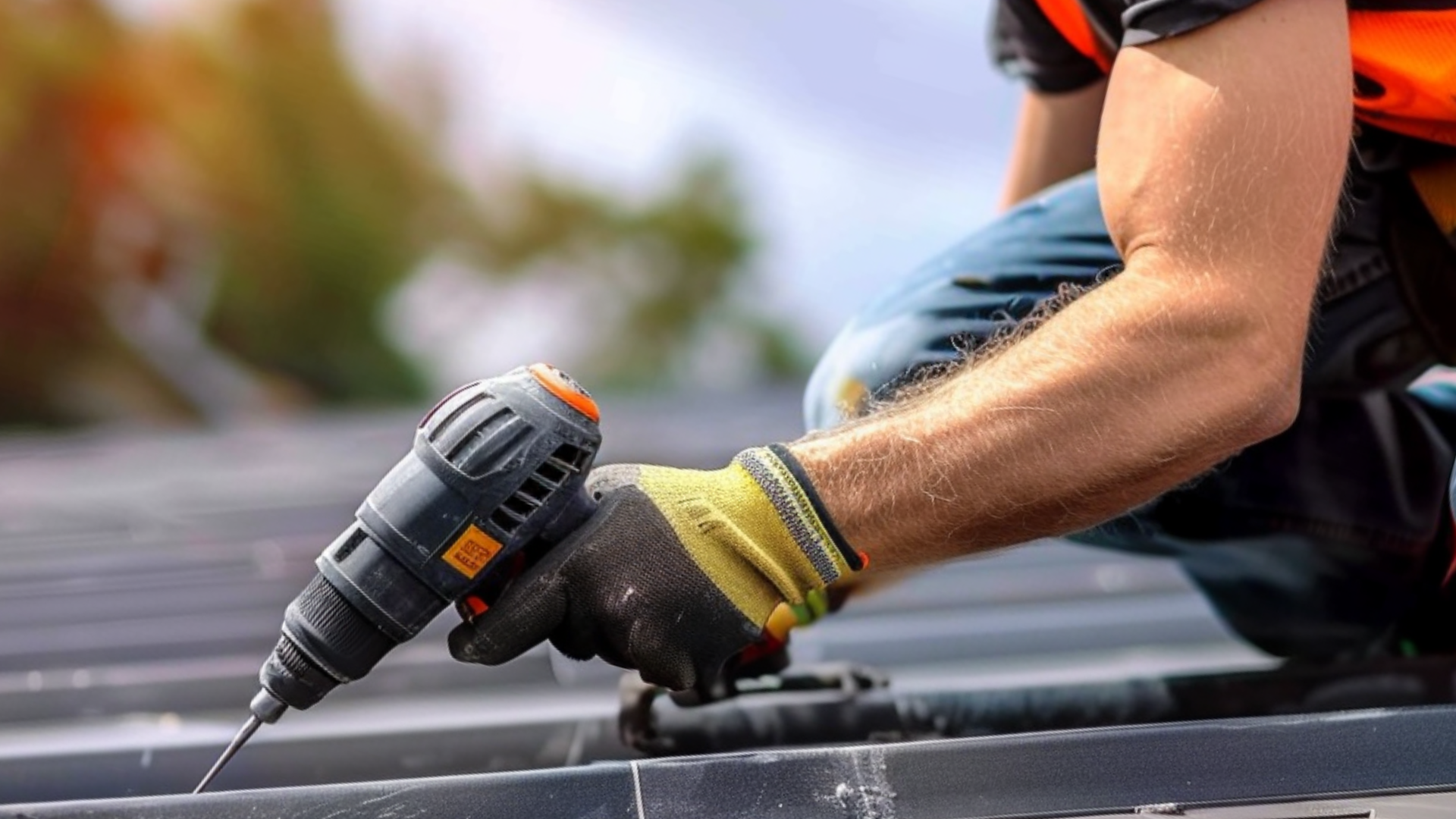 A roofer holding a screwdriver drill on top of the roof, nailing screws; he is wearing safety gloves.